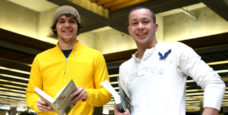 2 Students holding books in the library, main floor smiling at the camera
