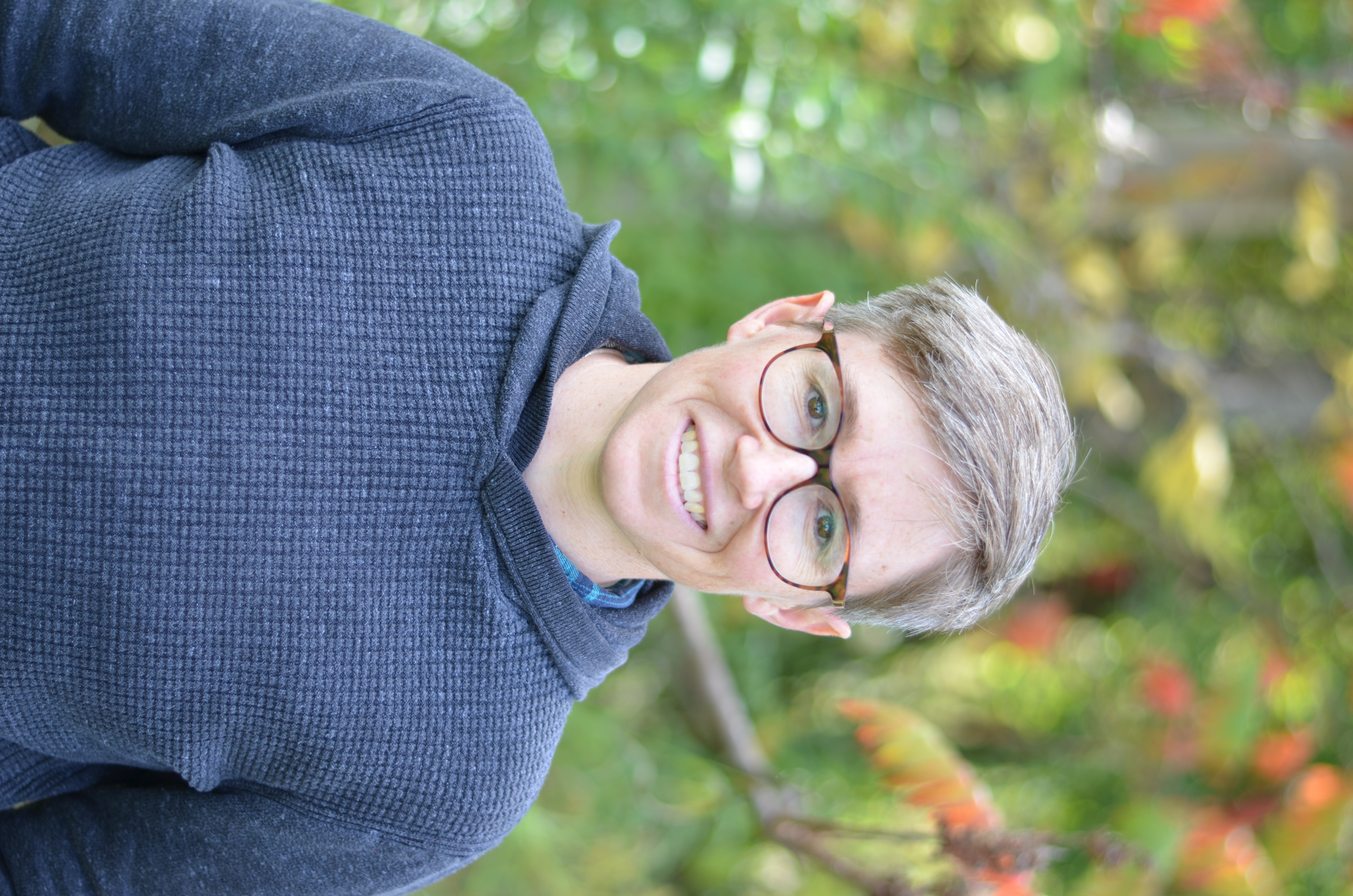 Sara Pieper in front of a group of sumac trees wearing glasses and a grey sweater, smiling at the camera.