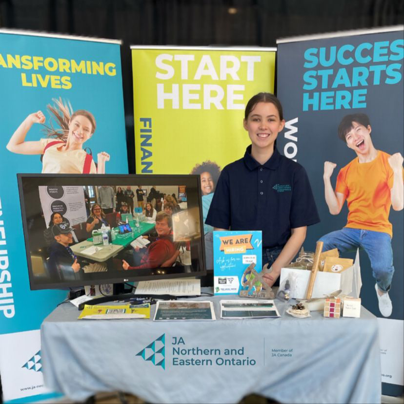 Smiling student standing behind an info table for JA Northern and Eastern Ontario with info banners behind them.