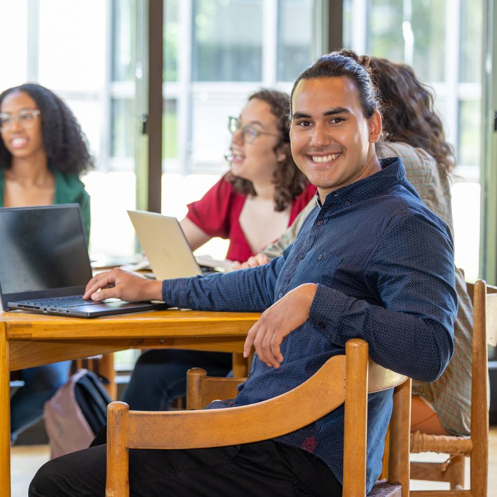 A man sitting in a chair at a table full of people. He has a hand on a laptop and is looking over the back of his seat and smiling at the camera.