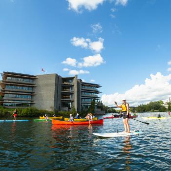 Students in canoes and on paddle boards on the Otonabee River boating past Bata Library.