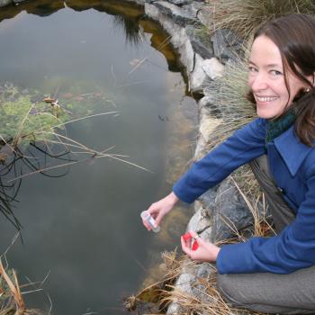 female kneeling down at water hole looking up smiling at camera with test tube in her hand taking a water sample