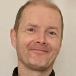 smiling man with pale skin and red hair outside on river with trees in background