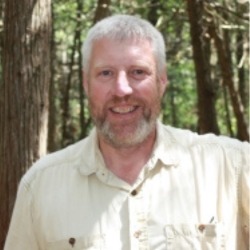 smiling man with pale skin, white hair and beard against a forest background