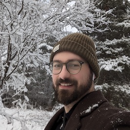 A person with a beard wearing a hat and eyeglasses against snowy tree background