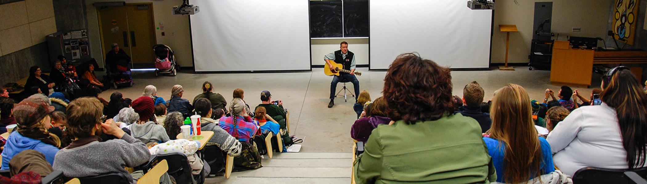 Ton Jackson sitting on a chair in a lecture hall playing guitar to an full audience