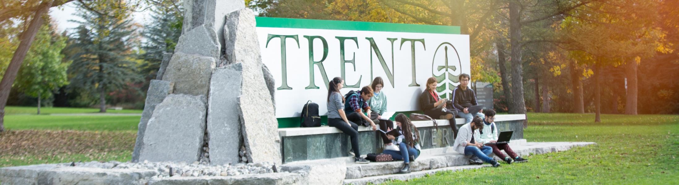 Students Sitting on the Trent sign at the entrance to Symons West Bank campus