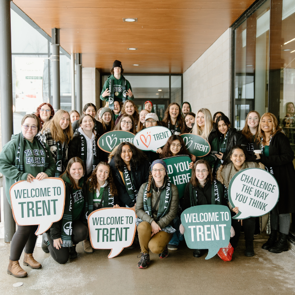 A group of Trent University students at Open House, holding welcoming signs. These signs include text such as, welcome to Trent, Challenge the way you think and I love Trent with a heart.