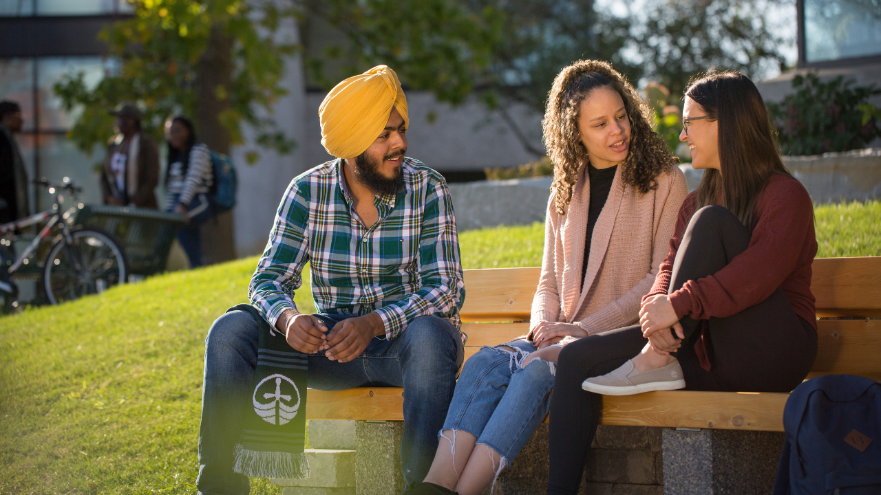 Three Trent University students sitting on a bench, talking to each other.