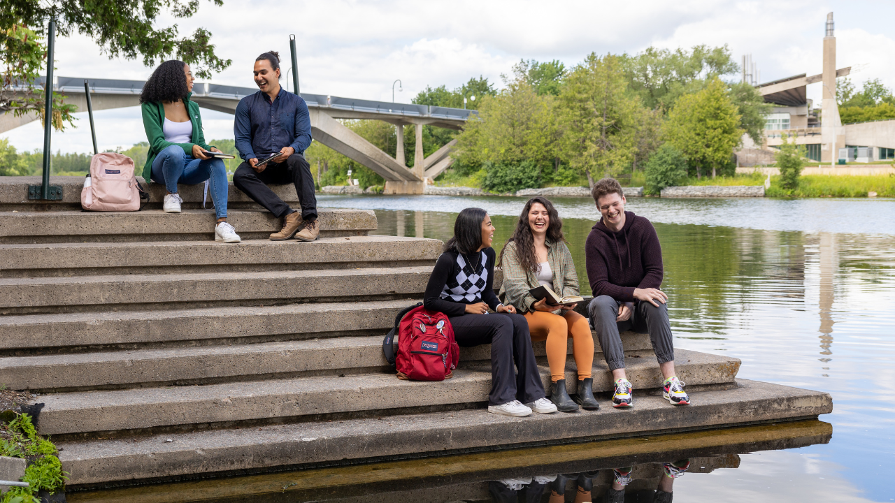 A group of five Trent students sitting outside of Bata library, with Otonabee river and Faryon bridge in the background.