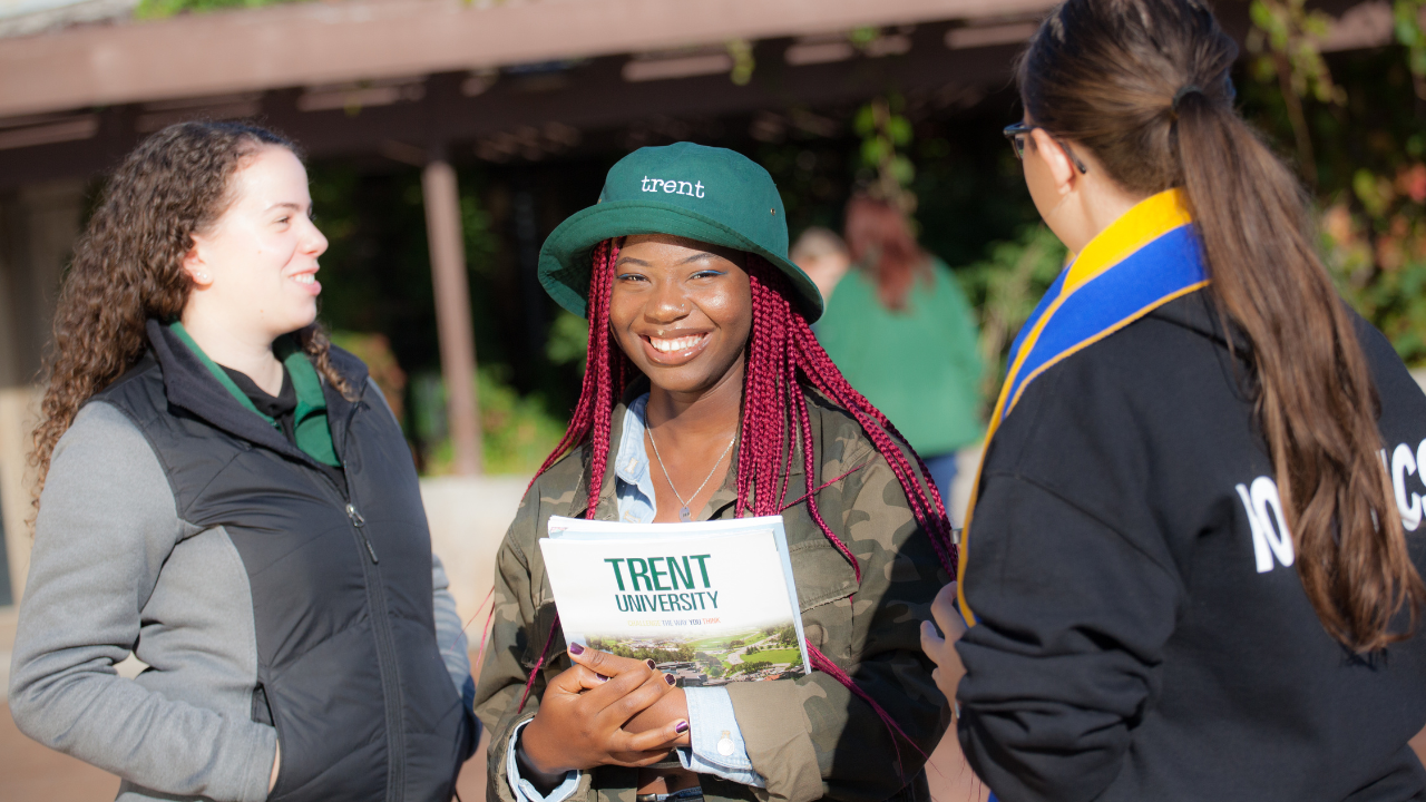 Three Trent University students outside together, with one student holding a Trent Viewbook and smiling at the camera.