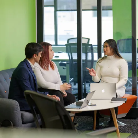 Three students sitting together at a table, in a office, with their laptops, talking to each other.