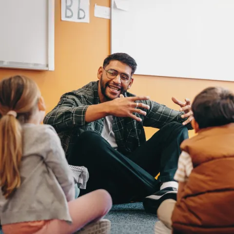 Image of a teacher sitting on the ground and talking with elementary school students.