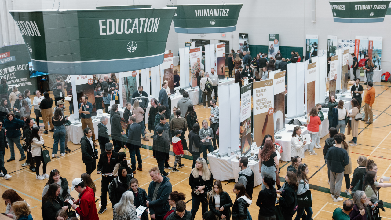 Image shows the Trent University gym during open house, there are signs to show programs, such as education and humanities. With crowds of students, families and staff talking.