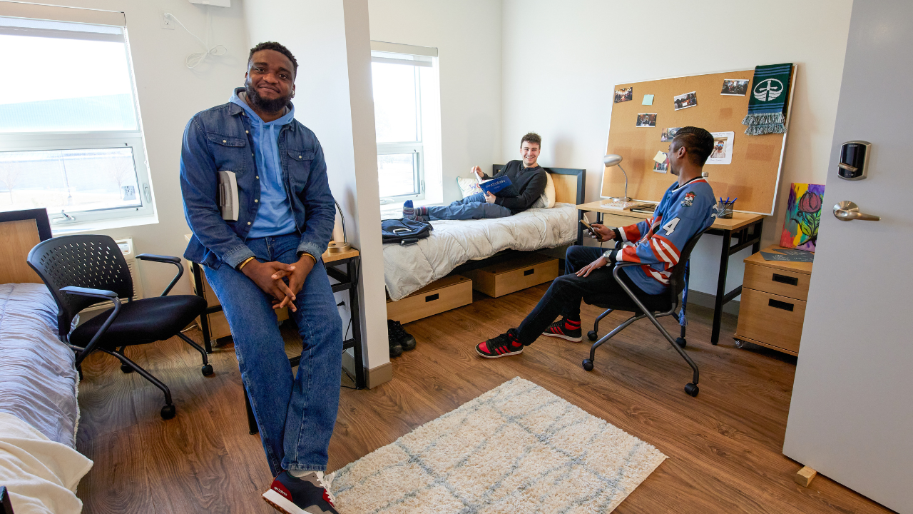 Trent Durham students in their dorm room, two are chatting on the bed while another student smiles at the camera.