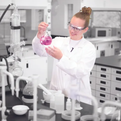 Trent University student in lab, holding up a chimerical glass beaker.