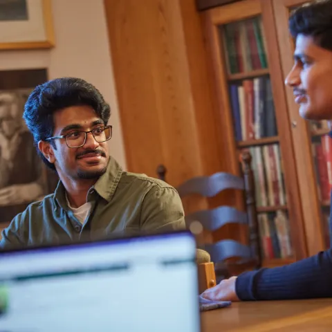 alt="Two students sitting at a table in Kerr House at Traill College, talking to each other, with an out of focus computer screen in the foreground.