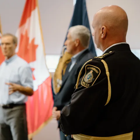 Image of three people, the person in the front is wearing a policing uniform, with a Peterborough Police badge in focus. In the background, the two mean are talking, with the Ontario and Canadian flag behind them