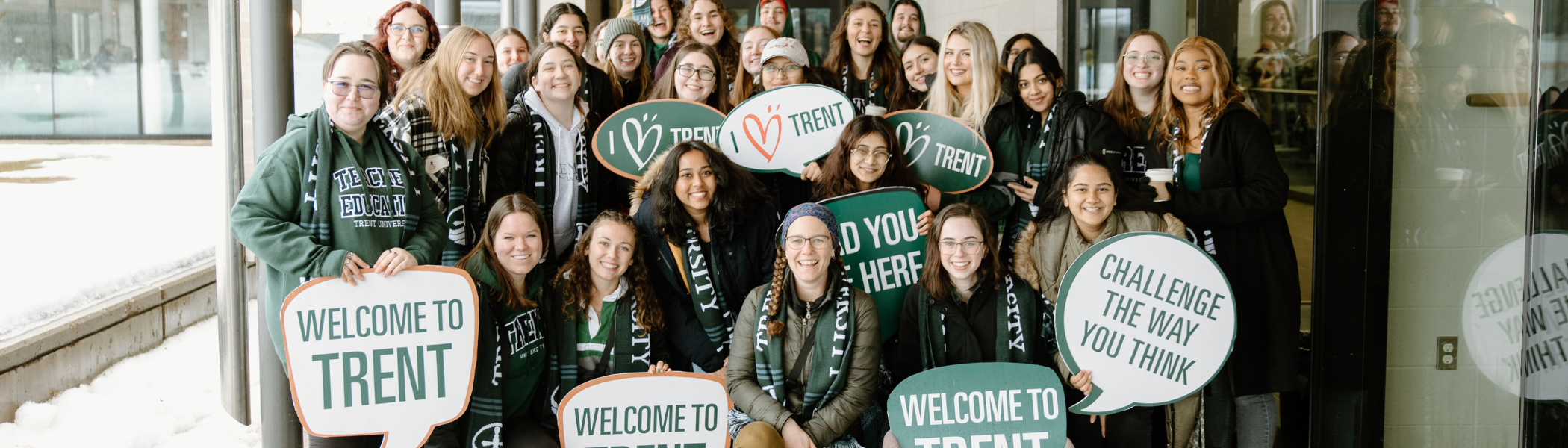 A group of Trent University students at Open House, holding welcoming signs. These signs include text such as, welcome to Trent, Challenge the way you think and I love Trent with a heart.  