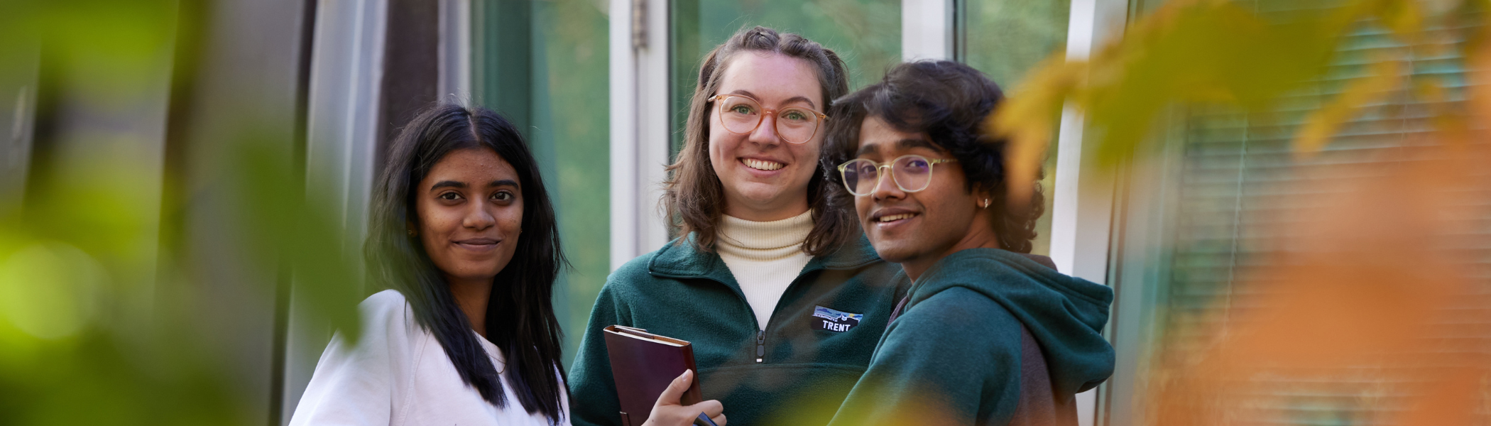 Three Trent University students smiling at the camera, with fall leaves around them. 