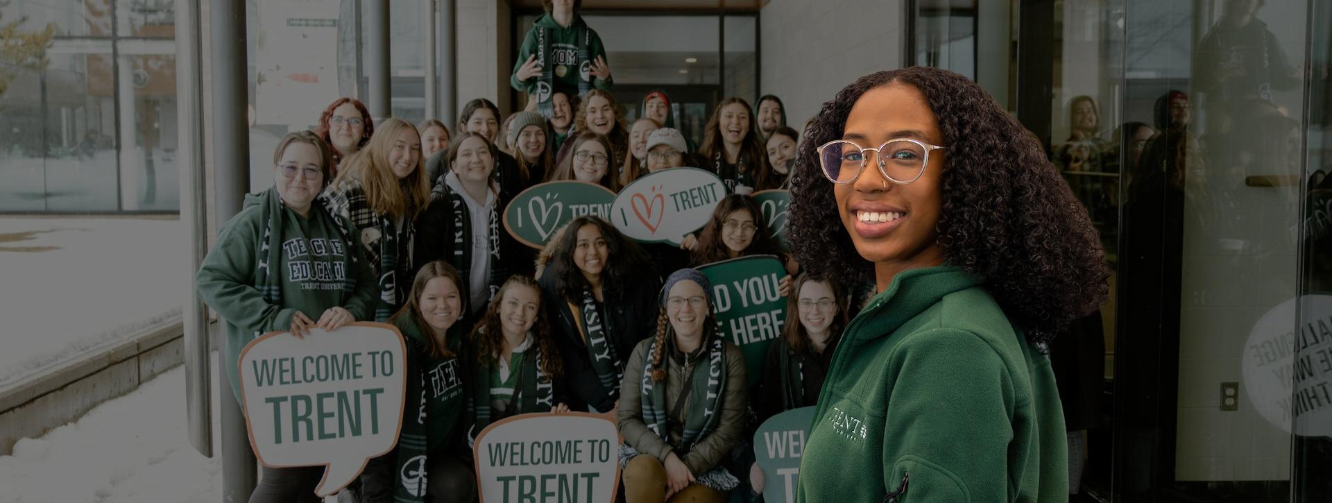 A group of diverse students standing together, smiling, and holding signs that say 'Welcome to Trent' and 'I ♥ Trent' in front of a building entrance with a snowy backdrop.