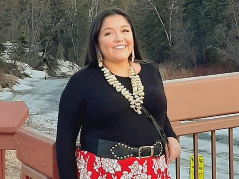 Smiling Indigenous woman with dark hair and shell necklace in front of frozen stream.