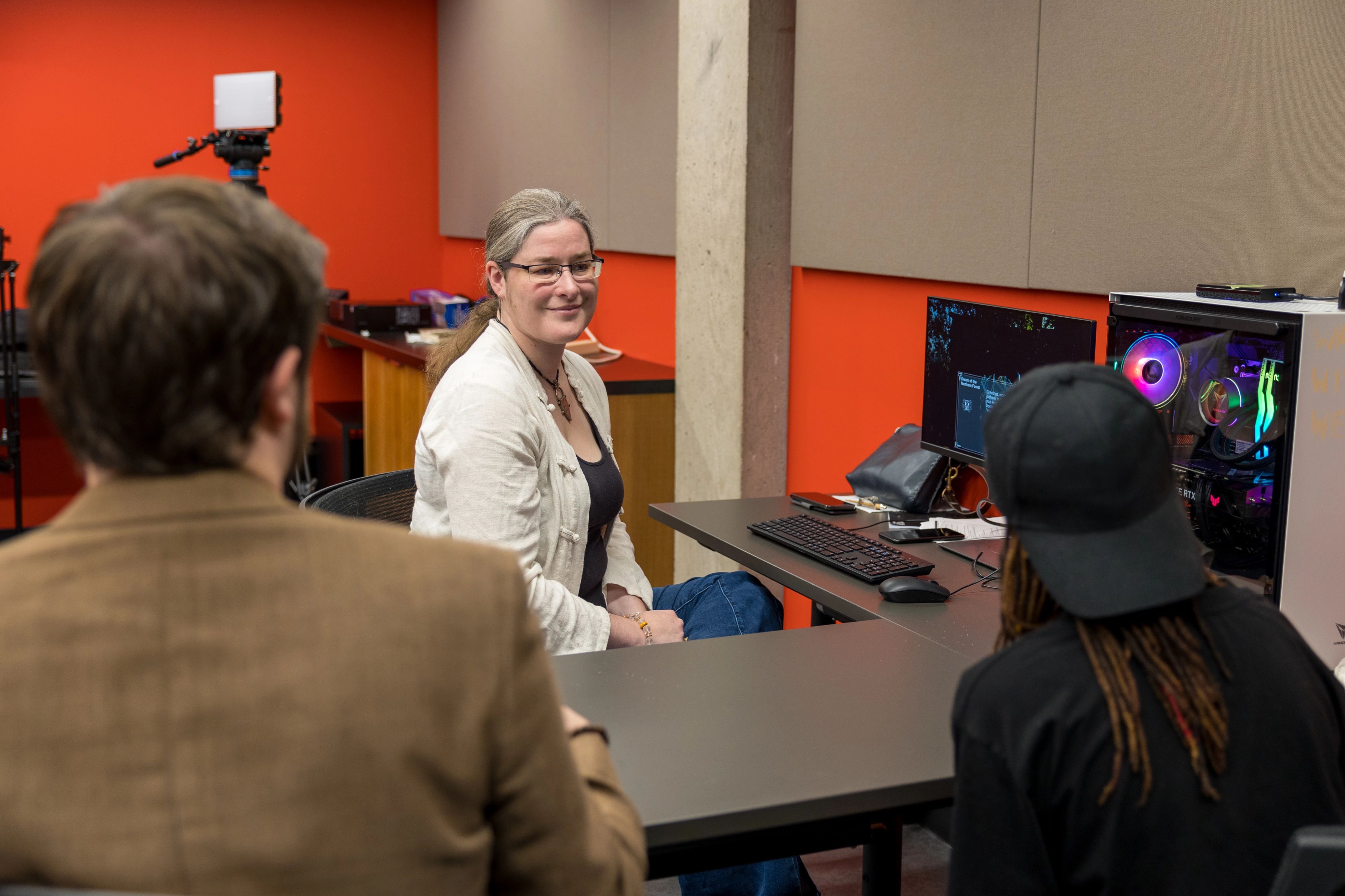 A female presenting women wearing white sitting at a computer with two students sitting across from her.