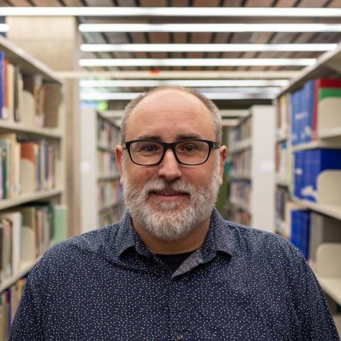 A man with glasses and a beard smiles in a library, wearing a blue shirt with small white patterns.