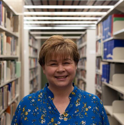A woman with short blonde hair smiles warmly in a library, wearing a blue floral blouse.