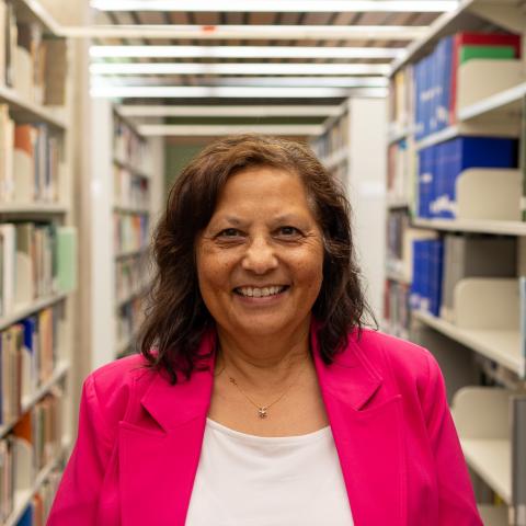 A cheerful woman wearing a bright pink jacket smiles broadly in a library aisle.