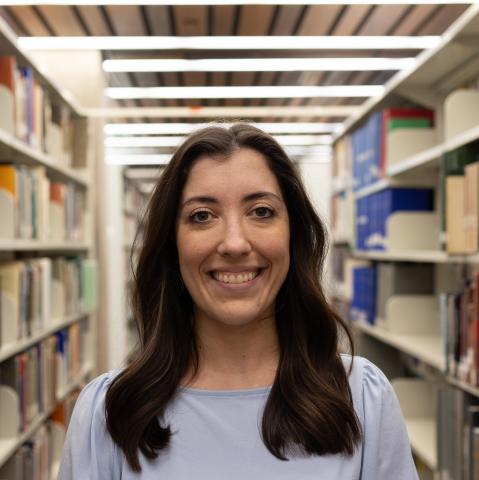 A woman with long dark hair, wearing a light blue shirt, smiles in an academic library.
