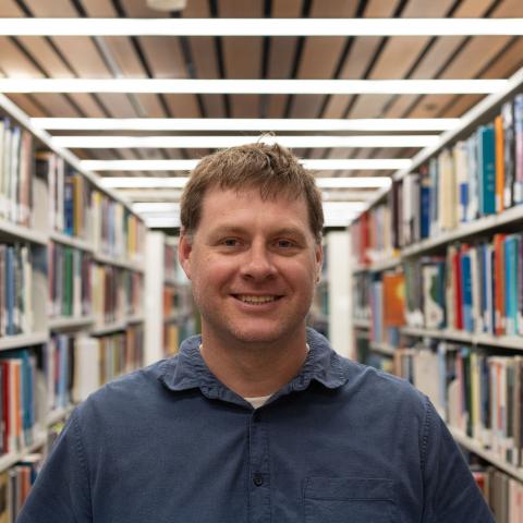 A person with short light brown hair is smiling while standing in a library. They are wearing a blue button-up shirt over a white t-shirt. Bookshelves filled with various books are visible in the background, creating a warm and academic atmosphere.