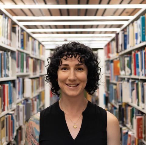 A person with curly black hair is smiling while standing in a library. They are wearing a black sleeveless top and a gold necklace with a small round pendant. Bookshelves filled with various books are visible in the background, creating a warm and academic atmosphere.