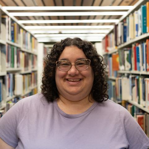 A person with curly dark hair and wearing glasses is smiling while standing in a library. They are dressed in a light purple short-sleeve top. Bookshelves filled with various books are visible in the background, creating a warm and academic atmosphere.