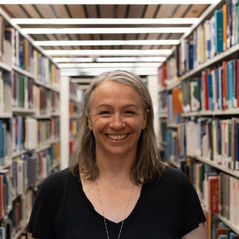 A person with shoulder-length gray hair is smiling while standing in a library. They are wearing a black short-sleeve top and a long necklace. Bookshelves filled with various books are visible in the background, creating a warm and academic atmosphere.