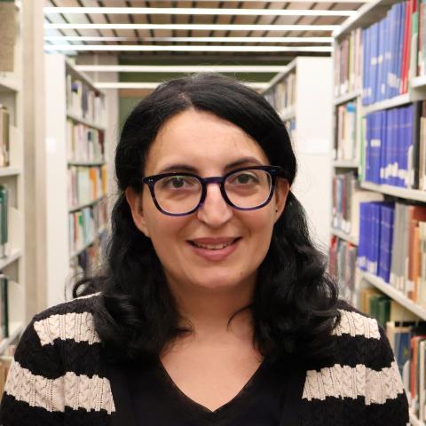 A woman with glasses and long dark hair, wearing a black and white striped cardigan, smiles in a library aisle lined with bookshelves.