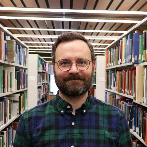 A man with glasses, a beard, and short brown hair, wearing a plaid shirt, smiles in a library aisle lined with bookshelves.