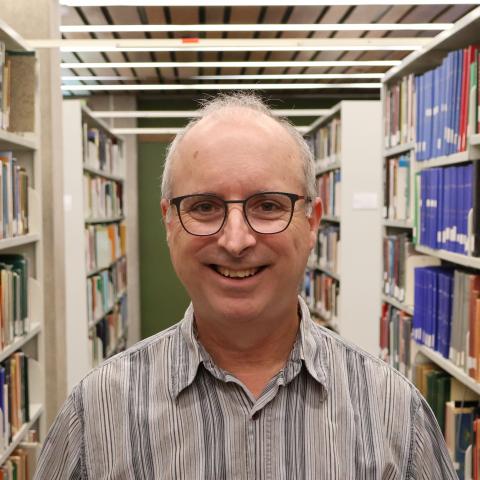 A man with glasses and short gray hair, wearing a striped shirt, smiles in a library aisle lined with bookshelves.