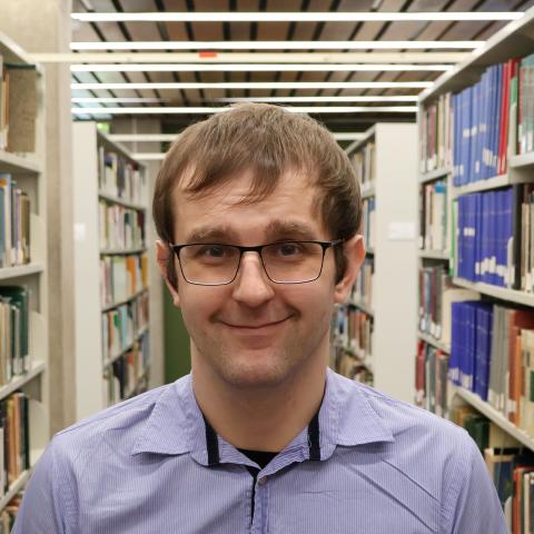 A man with glasses and short brown hair, wearing a light blue collared shirt, smiles in a library aisle lined with bookshelves.