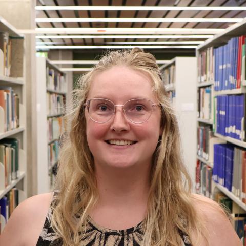 A woman with glasses and long blonde hair, wearing a sleeveless patterned top, smiles in a library aisle lined with bookshelves.