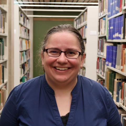 A woman with glasses and shoulder-length dark hair, wearing a blue blouse, smiles in a library aisle lined with bookshelves.