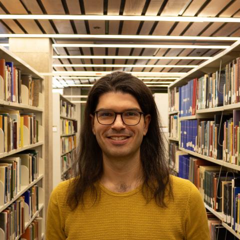 A smiling person wearing glasses and a yellow sweater stands in a brightly lit library aisle, surrounded by bookshelves.