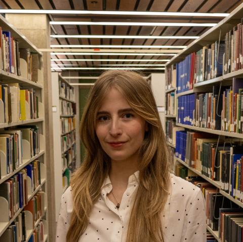 A smiling person with long blonde hair and a white polka-dotted shirt stands in a brightly lit library aisle, surrounded by bookshelves.