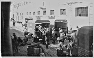 Black and white photograph of people in a cobblestone street, loading someone on a stretcher into an ambulance